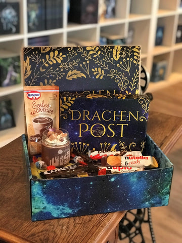 Das Produktbild zeigt vorn eine geöffnete Drachenpost in blauem Schuppen-Design. Man sieht in der Box ein in ein dunkellilafarbenes Drachenpost-Poster verpacktes Buch und zum Thema Schokolade passende Süßigkeiten sowie die Schoko-Kerze.